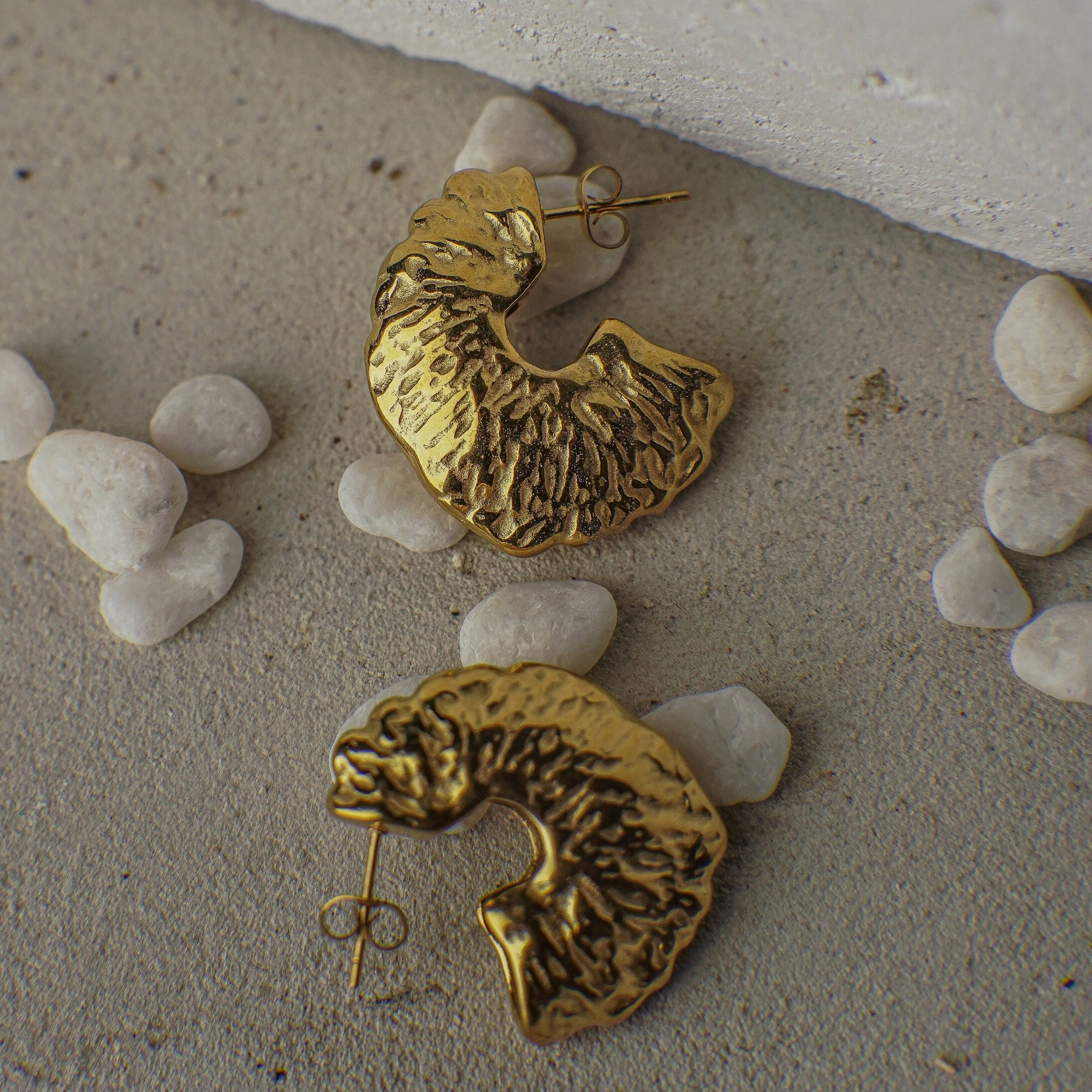Gold textured earrings on a stone surface with small stones around