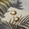A pair of gold hoop earrings placed on a white surface with a shadow of a plant leaf falling across them.