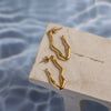 A pair of gold-colored drop earrings resting on a stone surface with water droplets in the background.