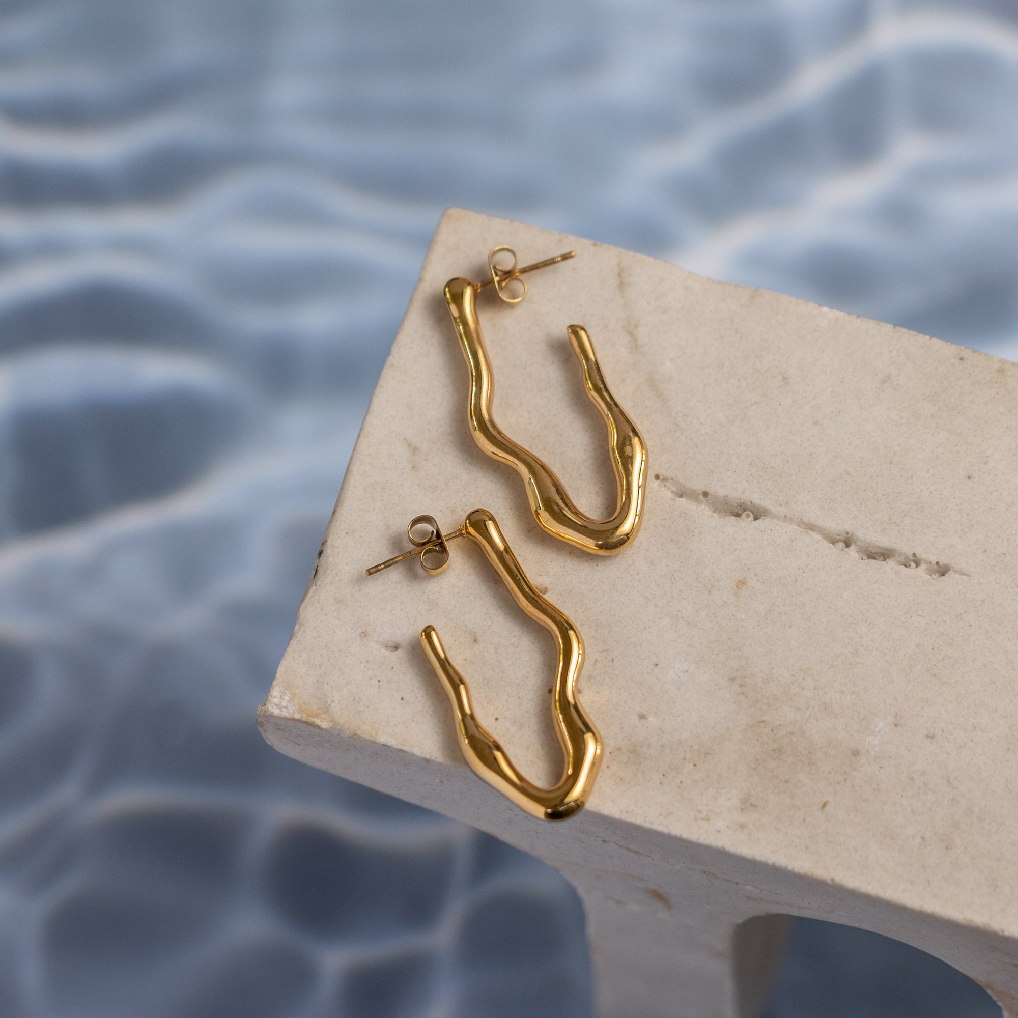 A pair of gold-colored drop earrings resting on a stone surface with water droplets in the background.