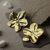 Gold flower-shaped earrings on a textured surface with pebbles.