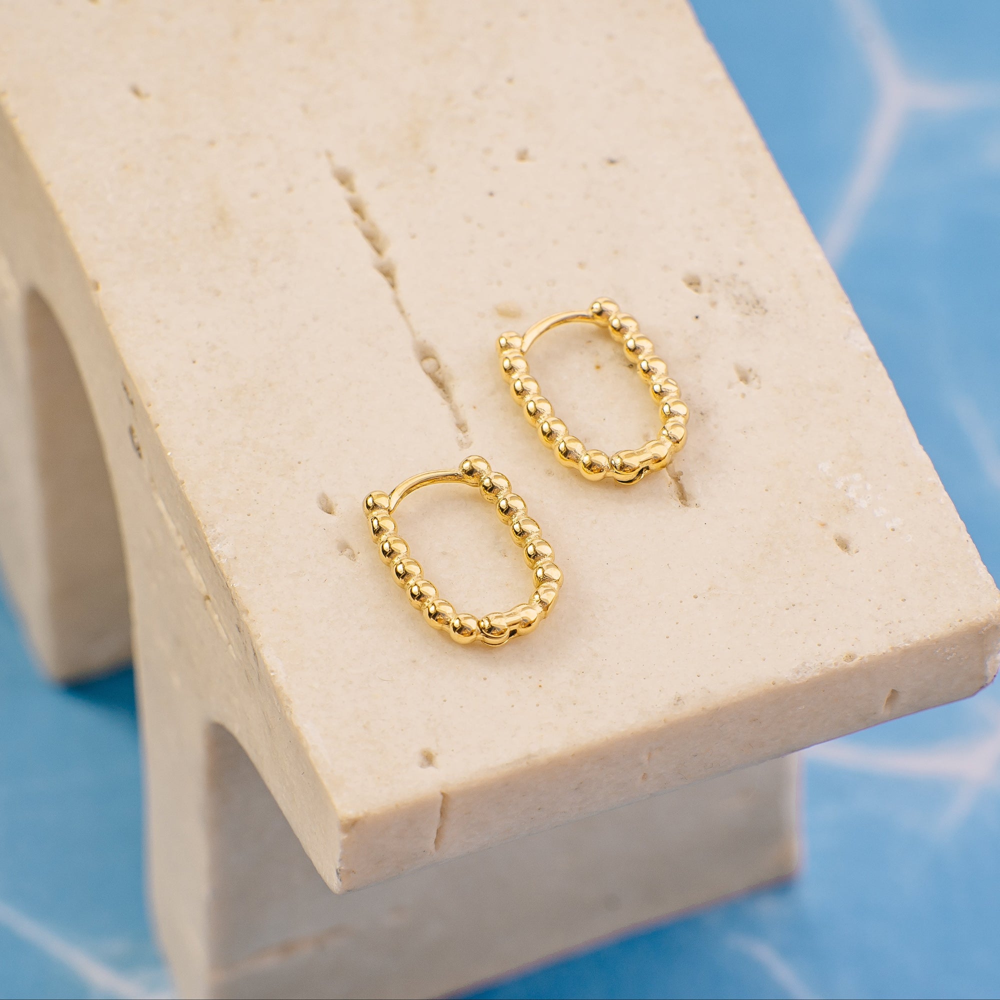 Gold hoop earrings on a beige stand with a blue geometric background