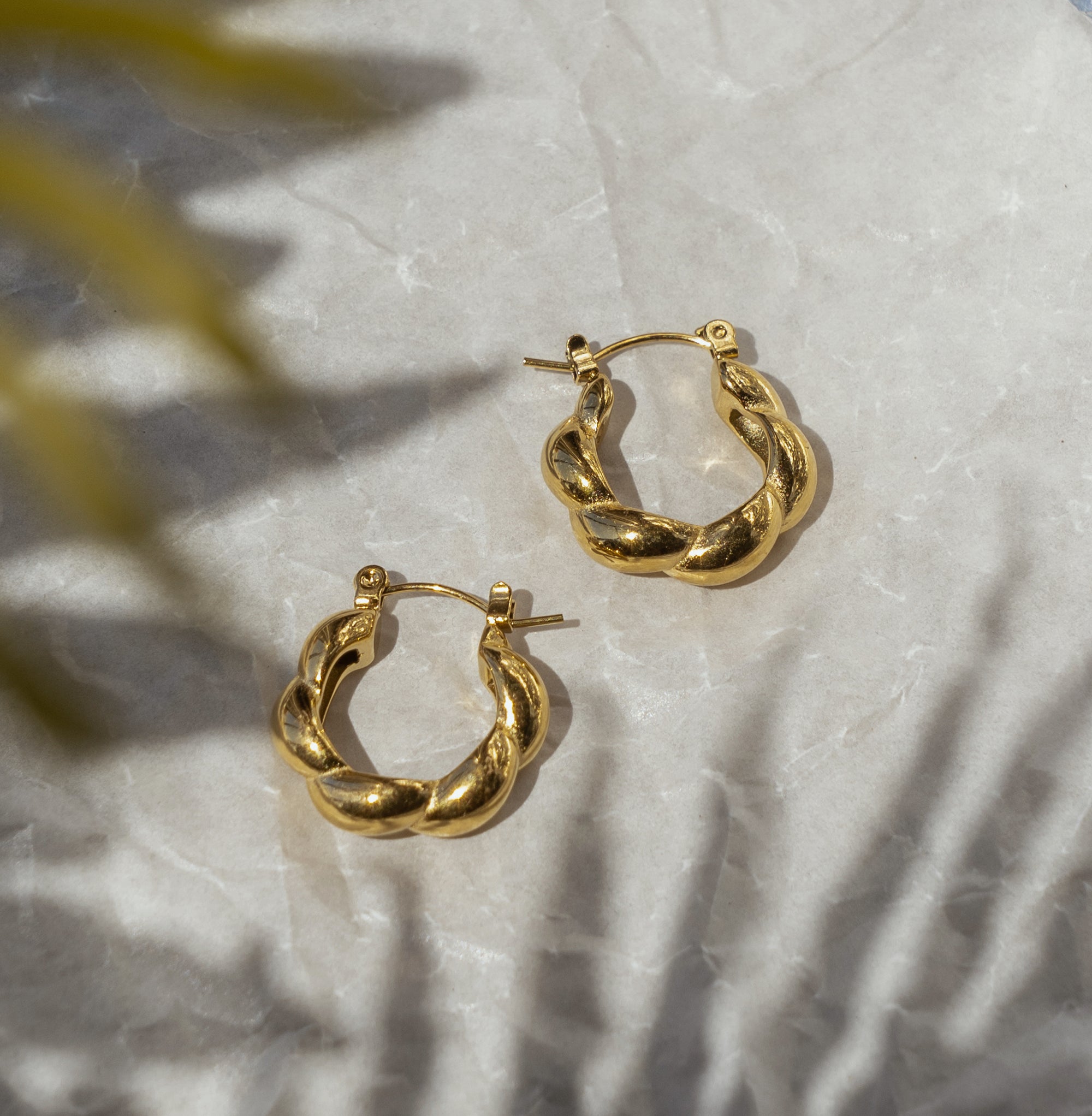 A pair of gold-colored textured hoop earrings placed on a textured surface with a plant leaf in the background.