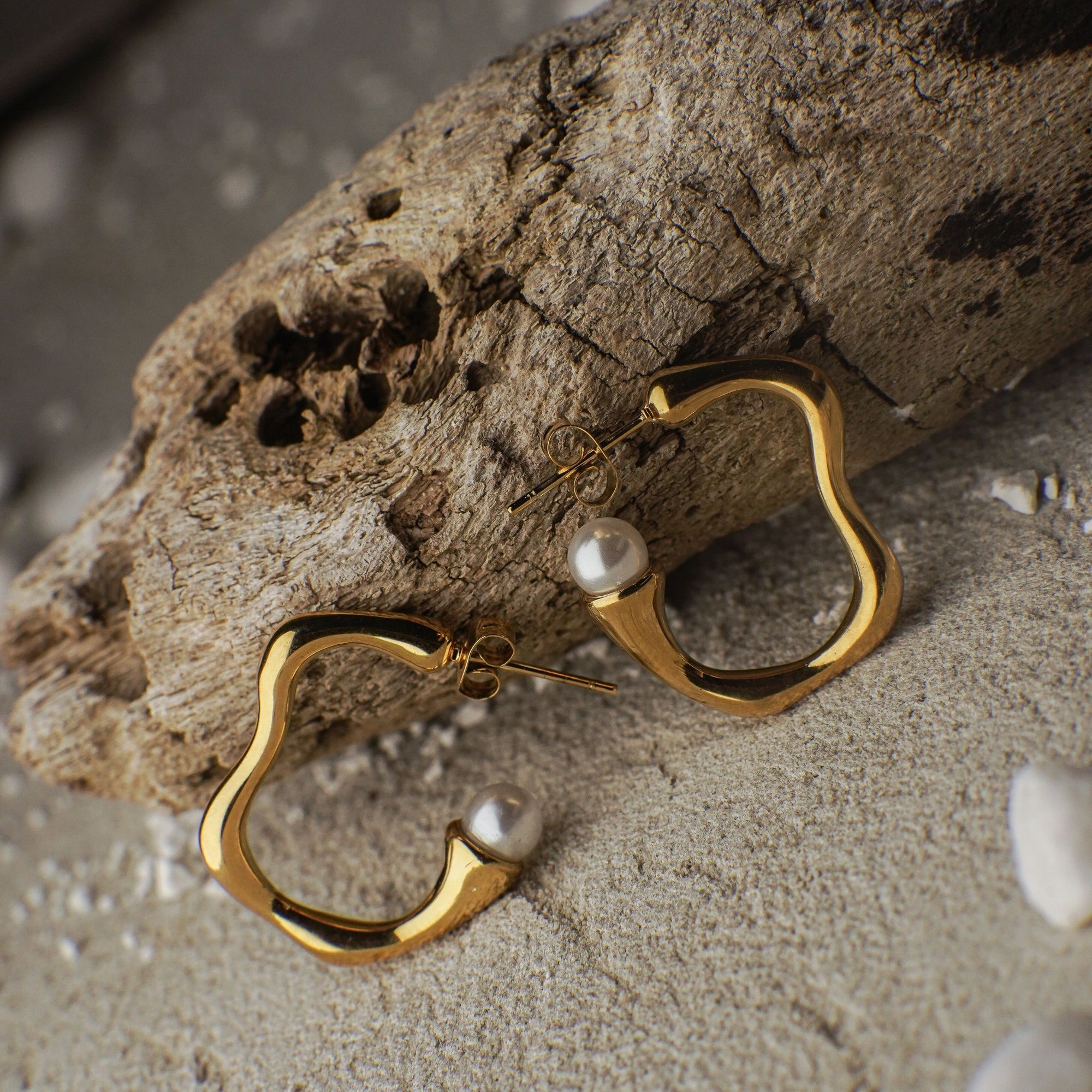 Gold hoop earrings with pearls on a sandy surface with driftwood.
