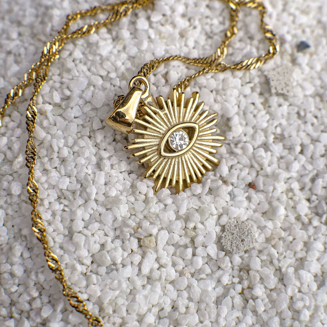 Gold necklace with a sunburst pendant on a sandy surface