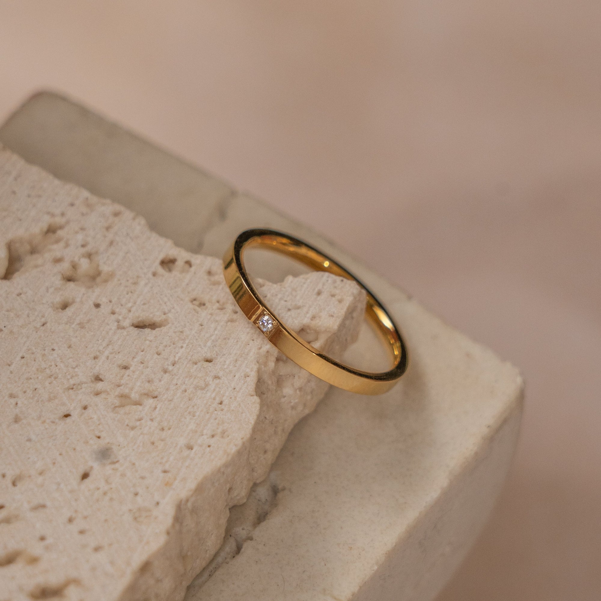 A gold-colored band ring with a zirconia centerpiece resting on a textured stone surface.