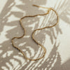 A gold-colored twisted chain necklace laid out on a flat surface with a shadow of a plant leaf pattern overhead.