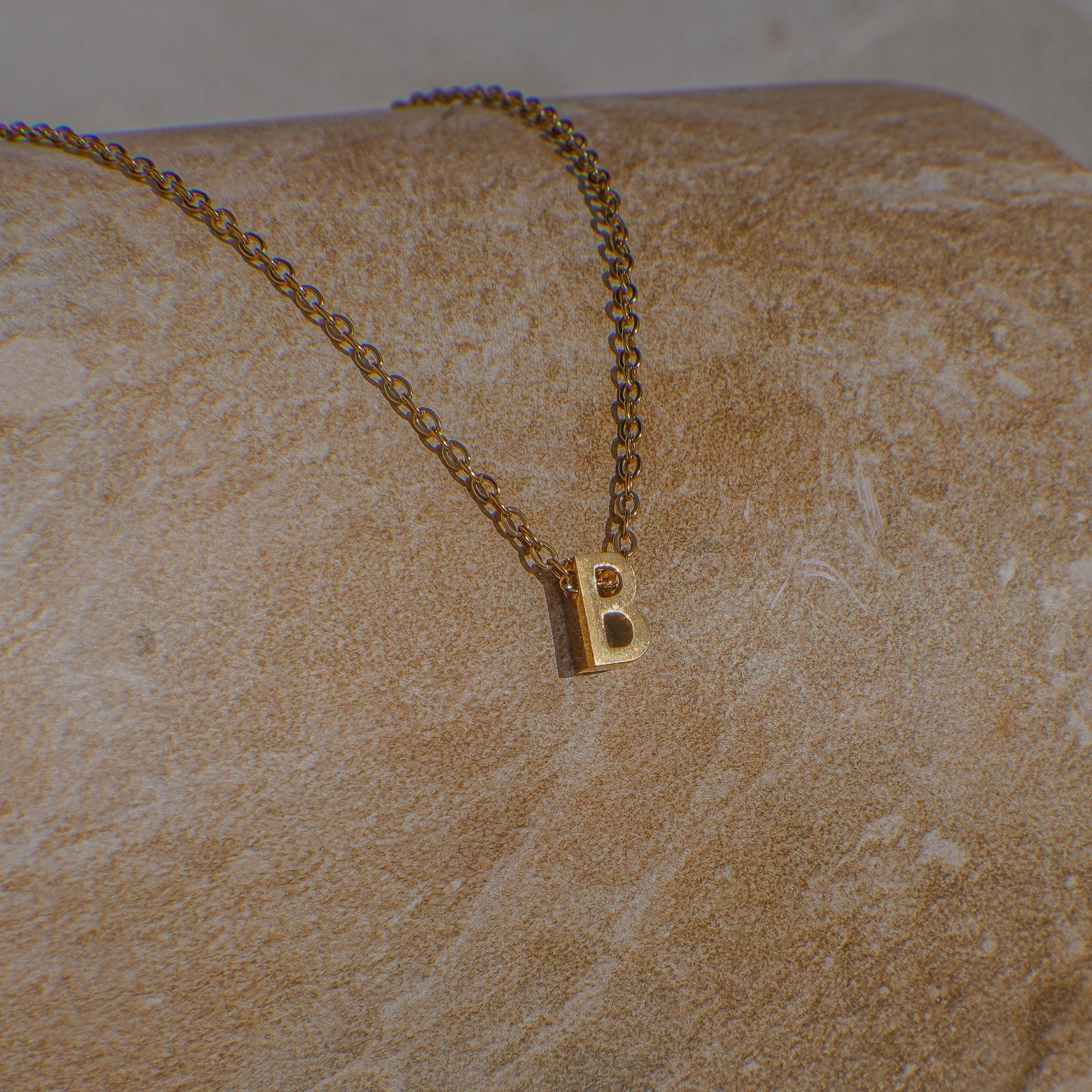 Gold necklace with a 'B' pendant on a beige stone surface