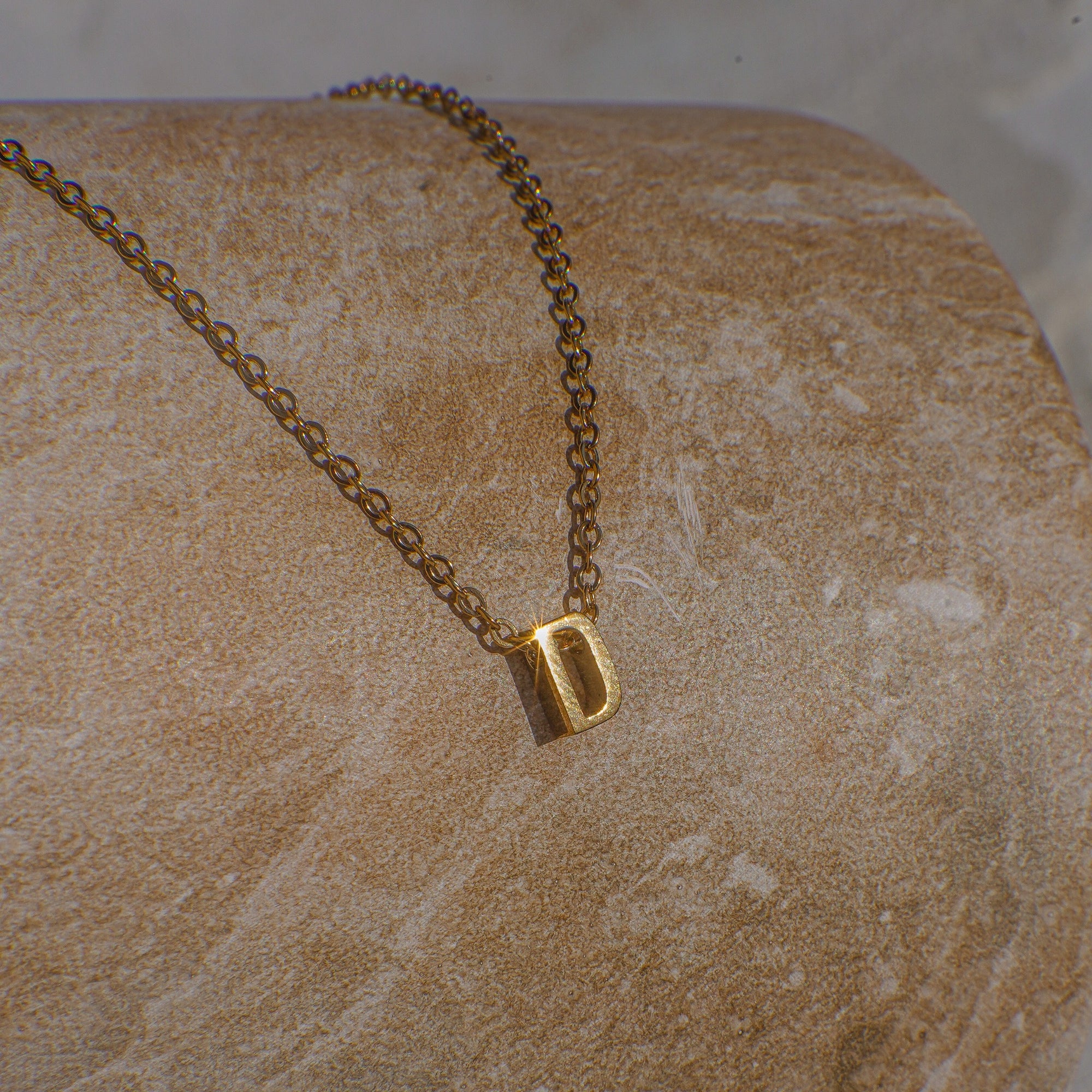 Gold necklace with a letter 'D' pendant on a beige stone surface