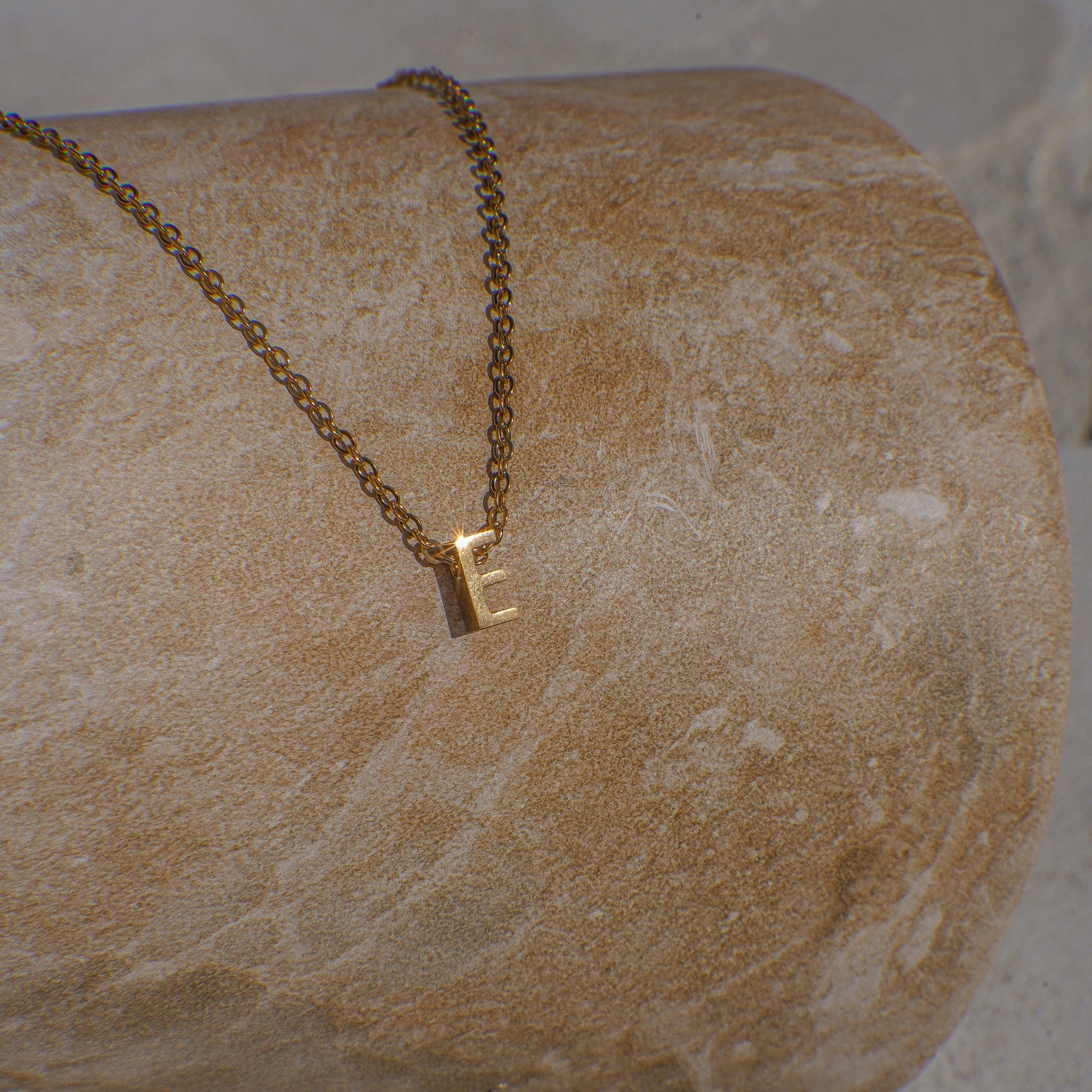 Gold necklace with a letter 'E' pendant on a textured stone surface