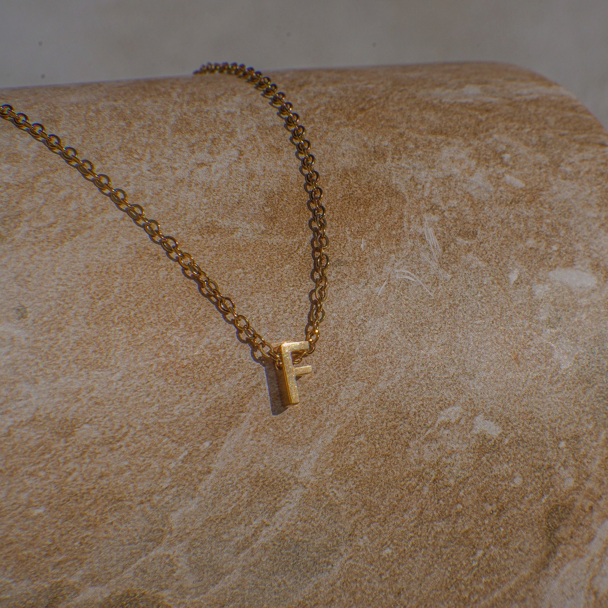 Gold necklace with a letter 'F' pendant on a textured beige surface