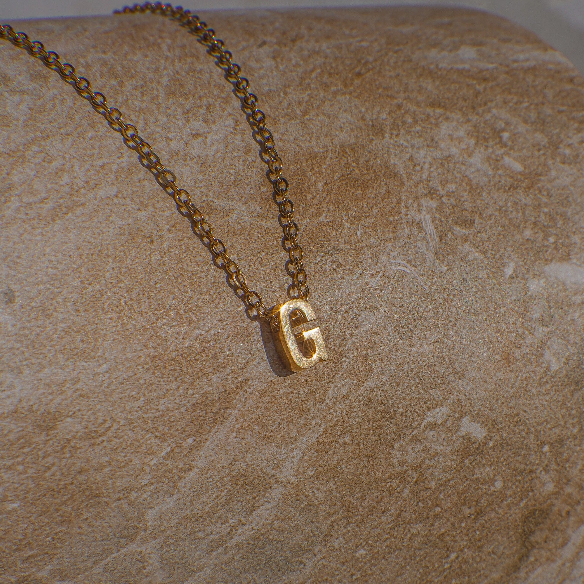 Gold necklace with a 'G' pendant on a beige stone surface