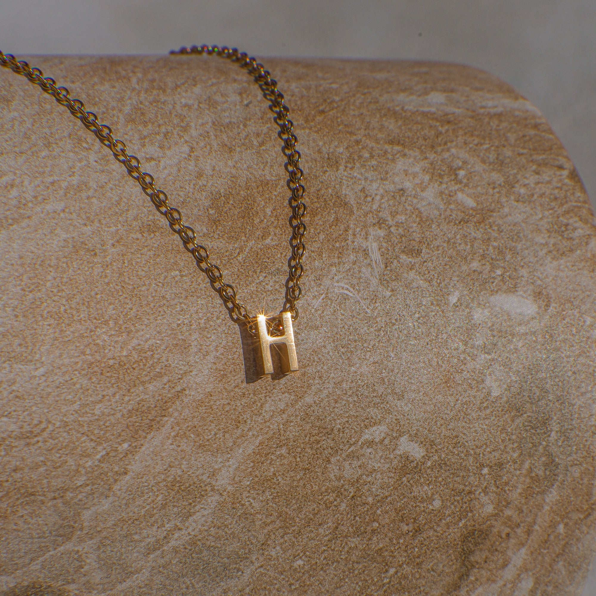 Gold necklace with a 'H' pendant on a stone surface