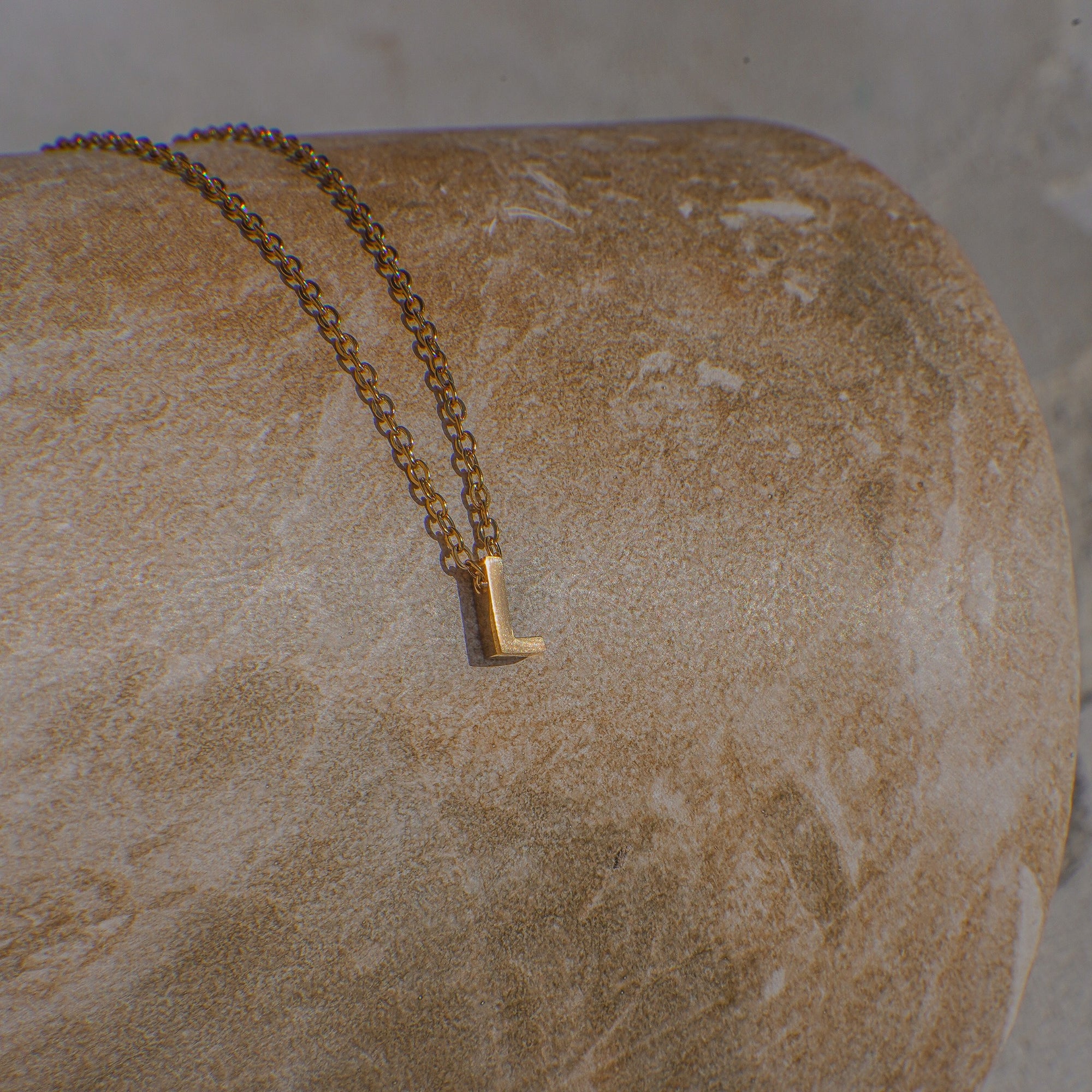 Gold necklace with a letter 'L' pendant on a textured stone surface