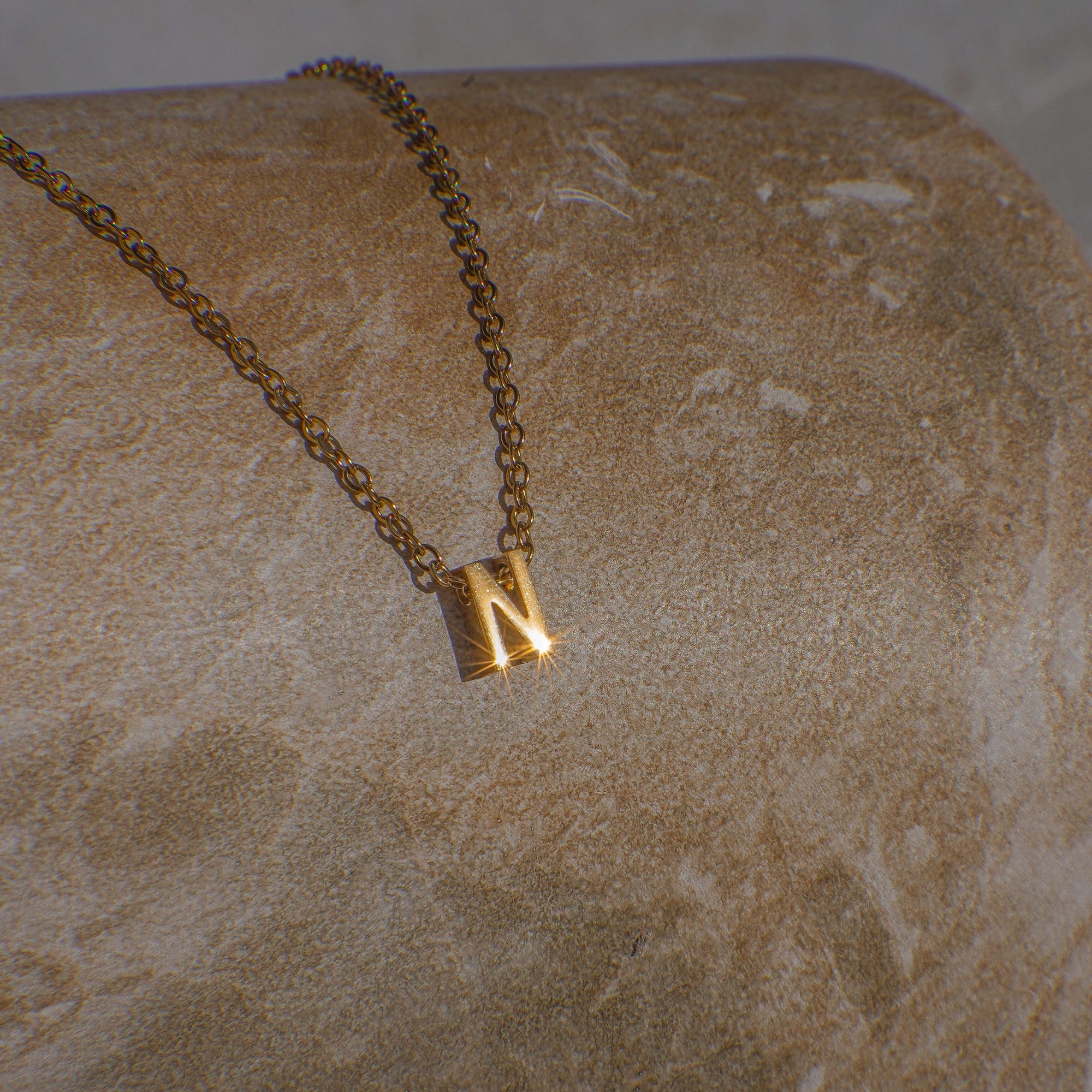Gold necklace with a 'N' pendant on a beige stone surface