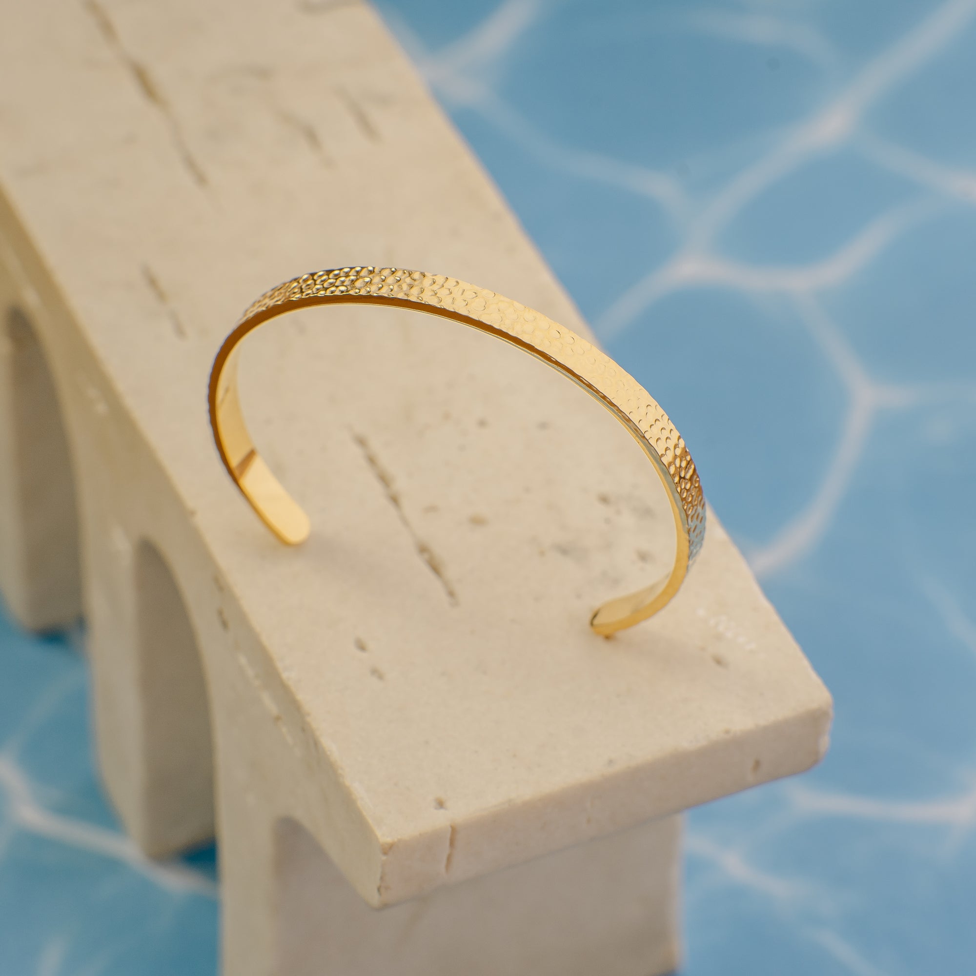 Gold bracelet on a white stand with a blue mosaic background
