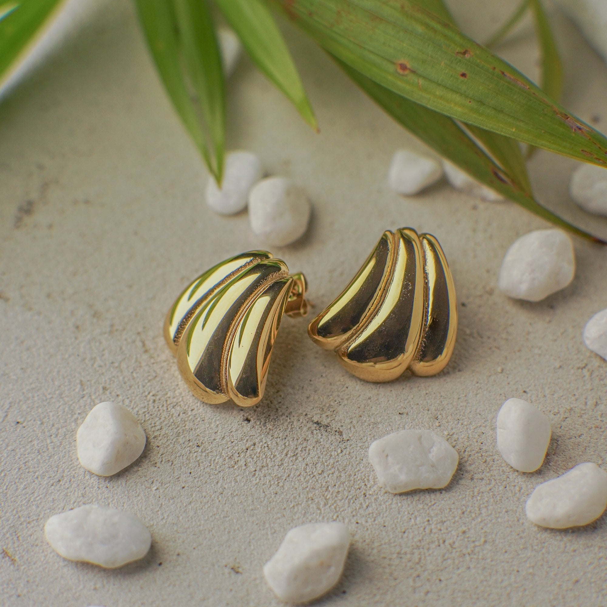 Pair of gold and black striped earrings on a sandy surface with pebbles and green leaves.
