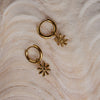 A pair of gold-colored hoop earrings with floral charm attachments, placed on a textured surface.