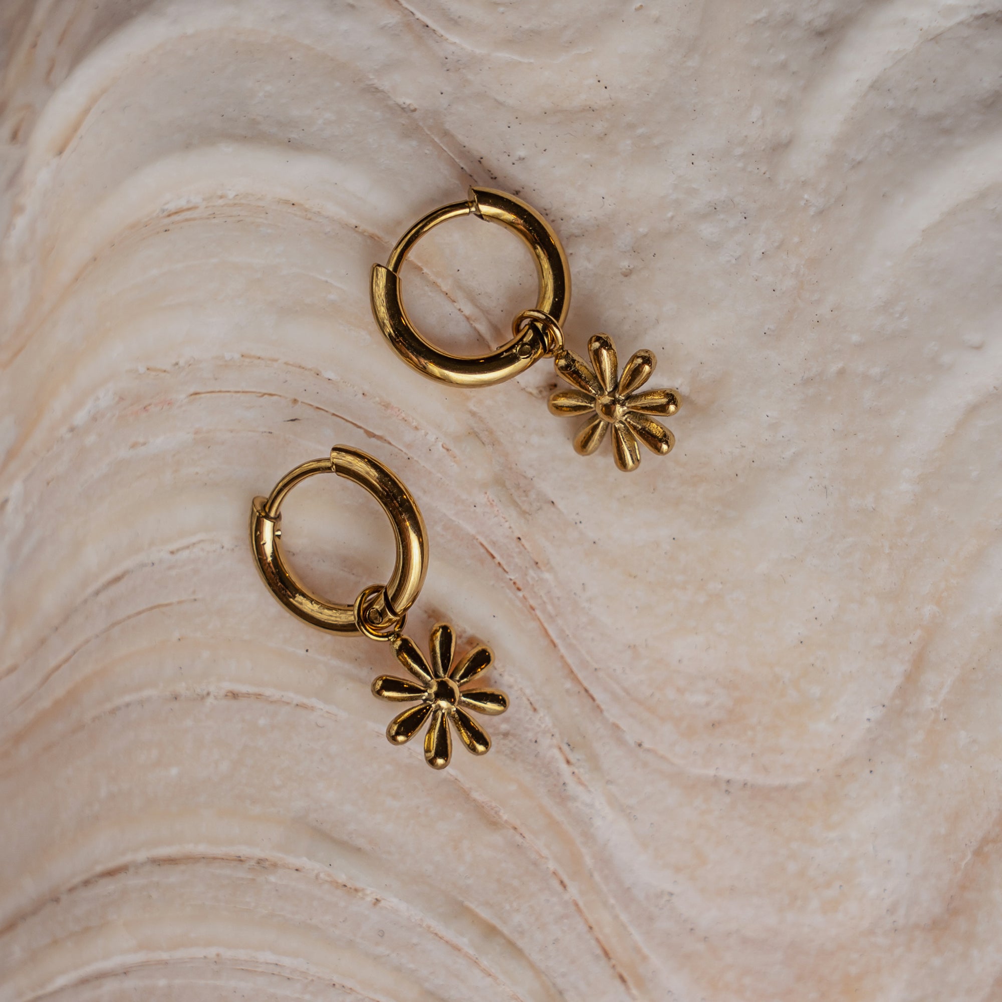 A pair of gold-colored hoop earrings with floral charm attachments, placed on a textured surface.