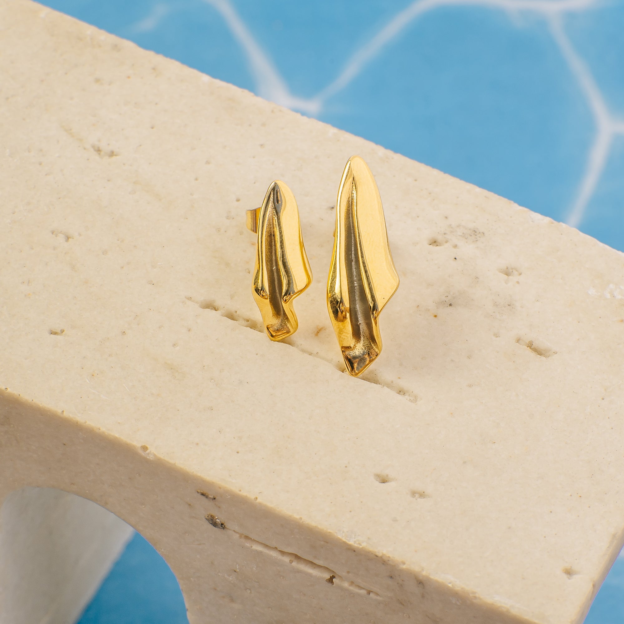 Gold earrings on a beige stone surface with a blue water background