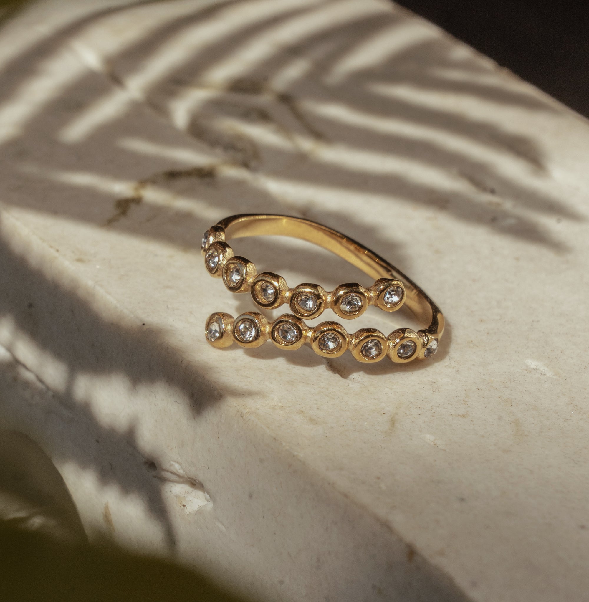 A gold ring with twisted zirconia bands and sparkling gems set in a ring, placed on a textured surface with a shadow falling over it.