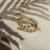 A gold-colored ring with multiple sparkling zirconia stones set around its circumference, placed on a marble surface with palm leaf shadows casting over it.