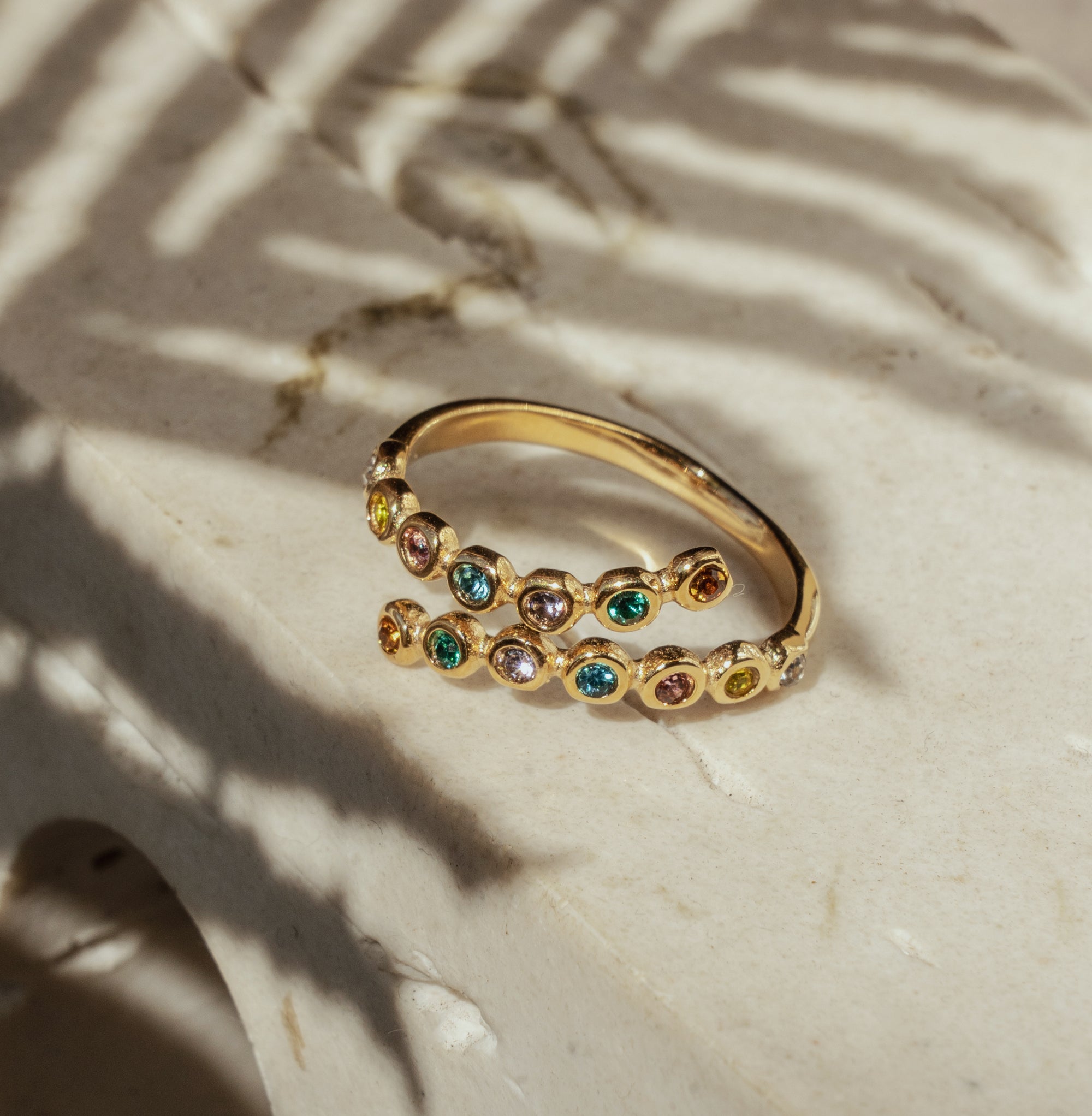A gold-colored ring with multiple sparkling zirconia stones set around its circumference, placed on a marble surface with palm leaf shadows casting over it.