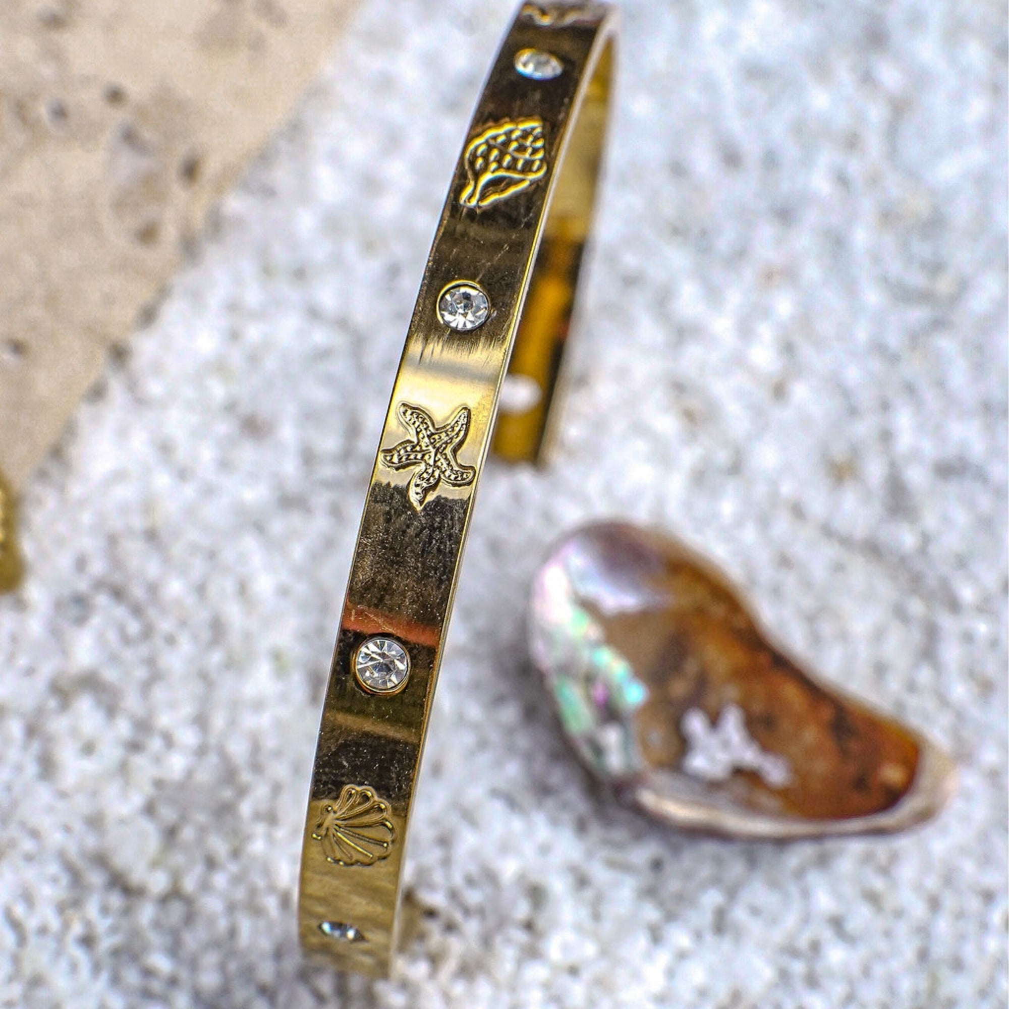 Gold bracelet with shell and starfish designs on a sandy background