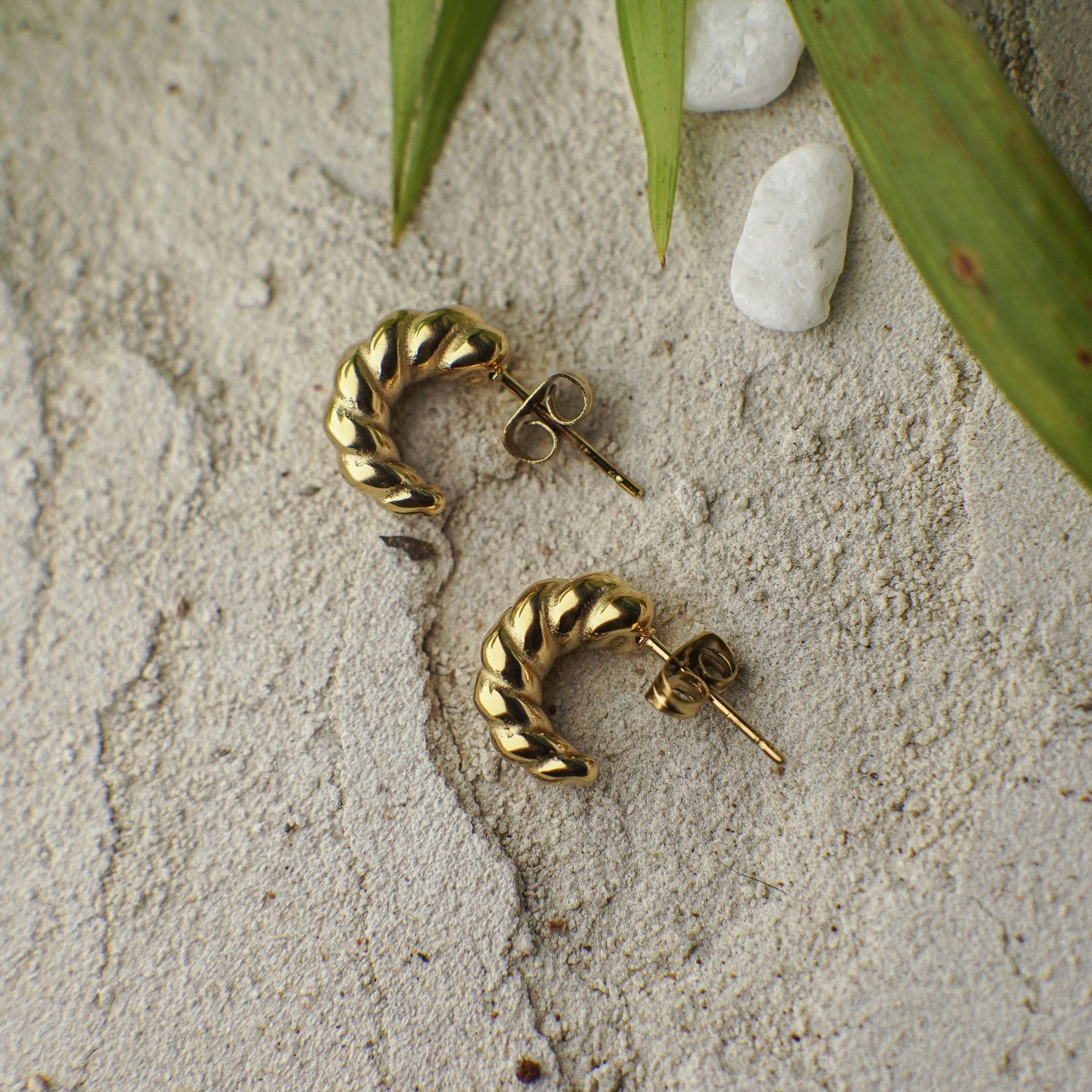 Gold hoop earrings on a textured surface with leaves and stones in the background