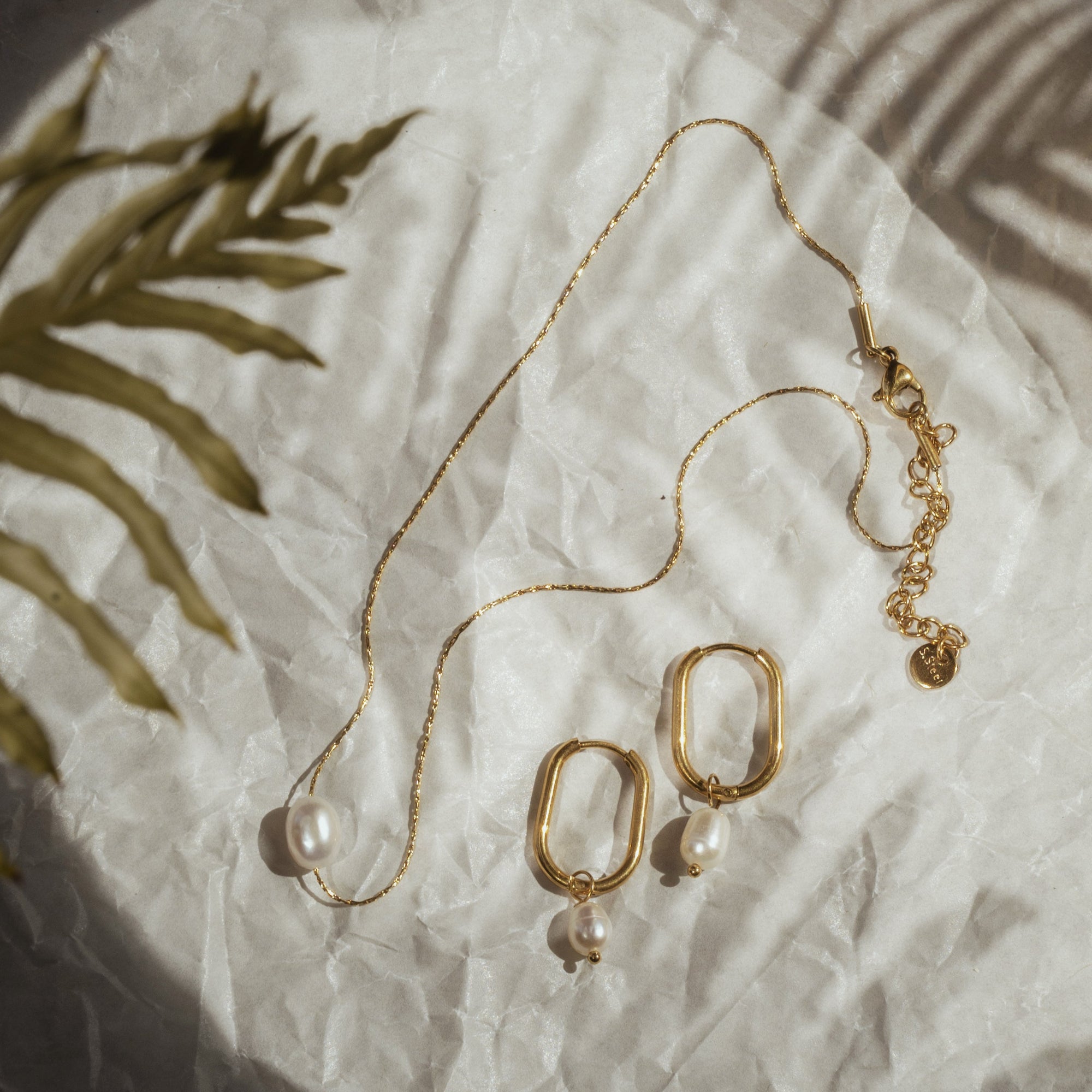 A gold-plated pearl necklace with a single pearl pendant, displayed on a white surface with a palm leaf in the background.