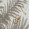 A gold-plated pearl necklace with a single pearl pendant and a pair of earrings with same features, displayed on a white surface with a palm leaf in the background.