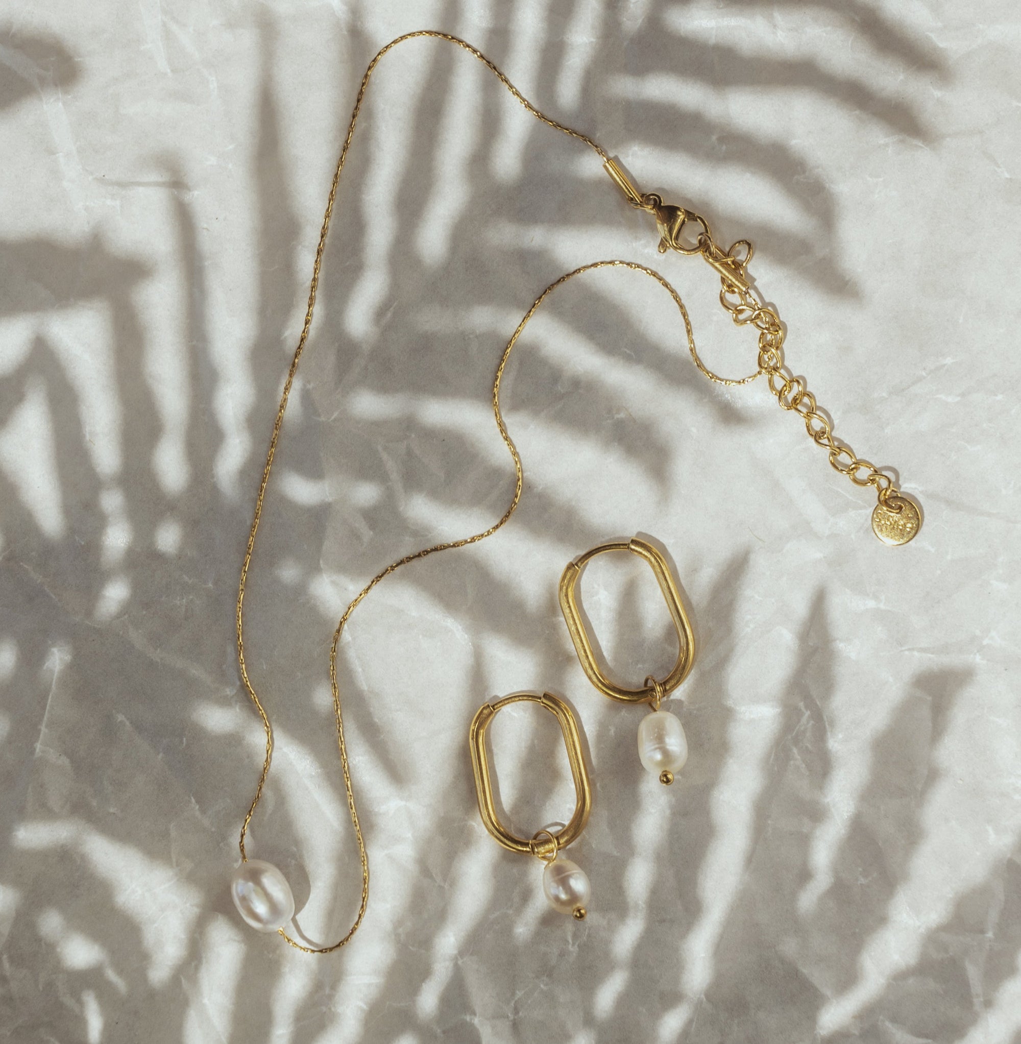 A gold-plated pearl necklace with a single pearl pendant and a pair of earrings with same features, displayed on a white surface with a palm leaf in the background.