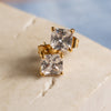 A pair of gold-colored earrings with square zirconia studs, displayed on a textured surface.