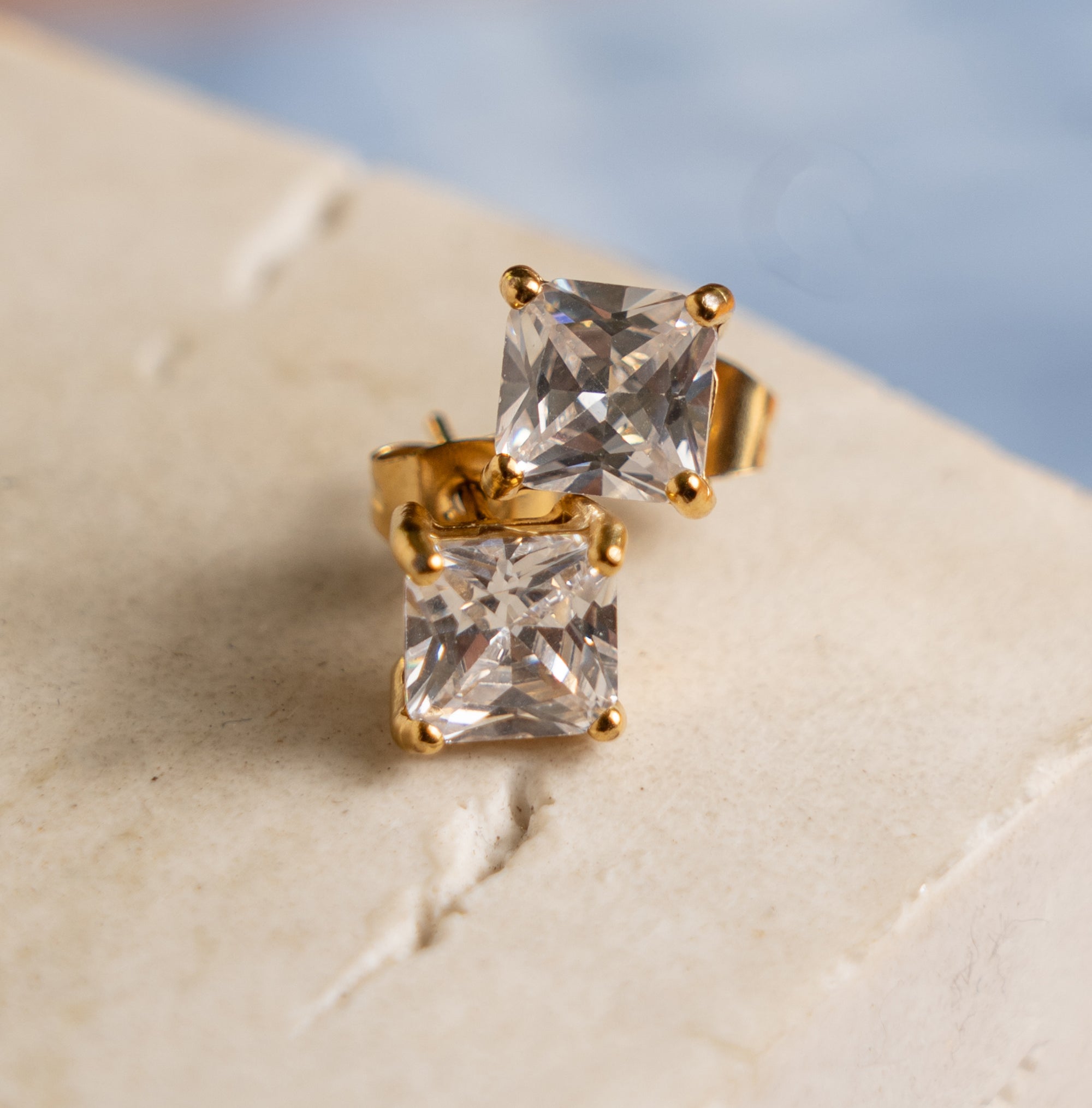 A pair of gold-colored earrings with square zirconia studs, displayed on a textured surface.