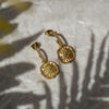 A pair of gold-colored, sun-inspired earrings with intricate details, placed on a textured surface with a shadow of a plant leaf.