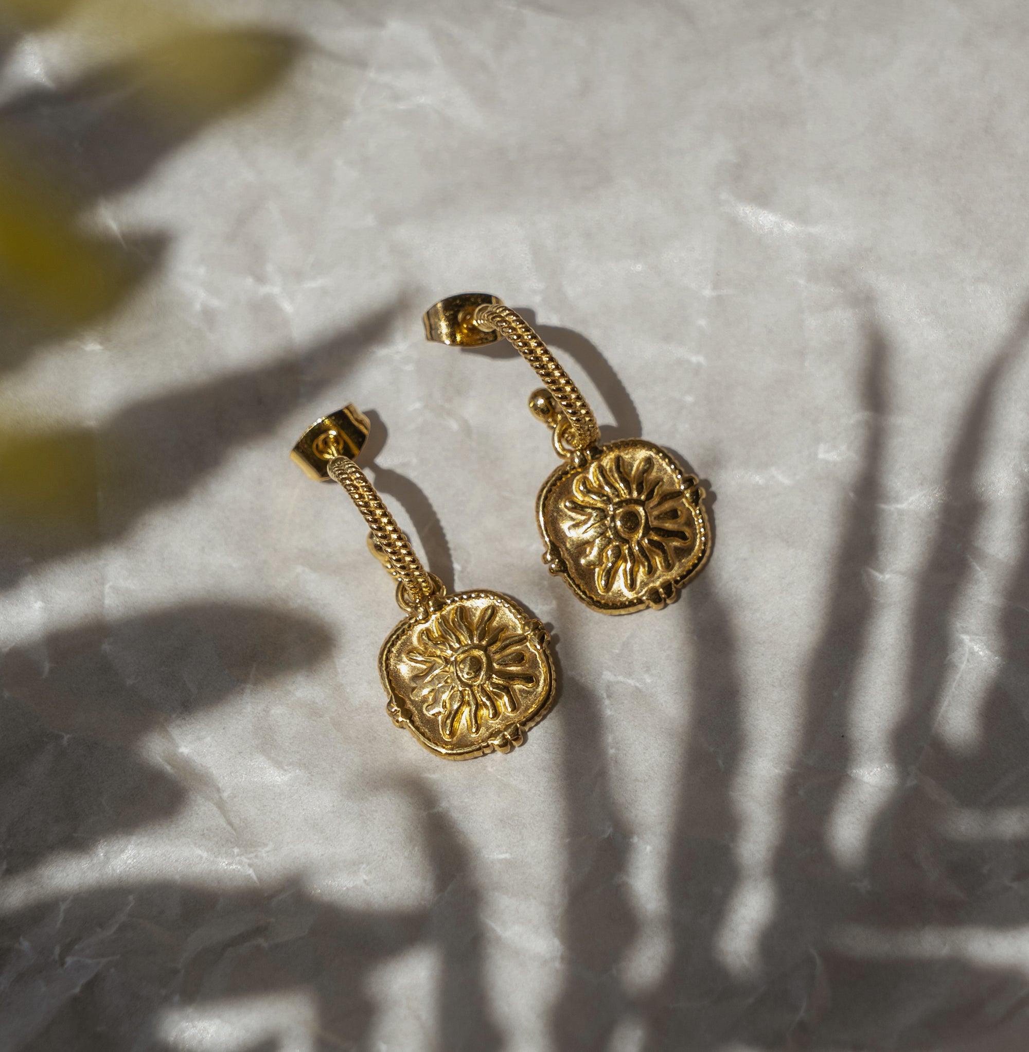 A pair of gold-colored, sun-inspired earrings with intricate details, placed on a textured surface with a shadow of a plant leaf.