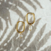 A pair of gold-colored hoop earrings with white pearls attached to them, displayed on a textured surface.