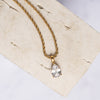 A gold-colored necklace with a clear teard-shaped zirconia pendant displayed on a beige background.