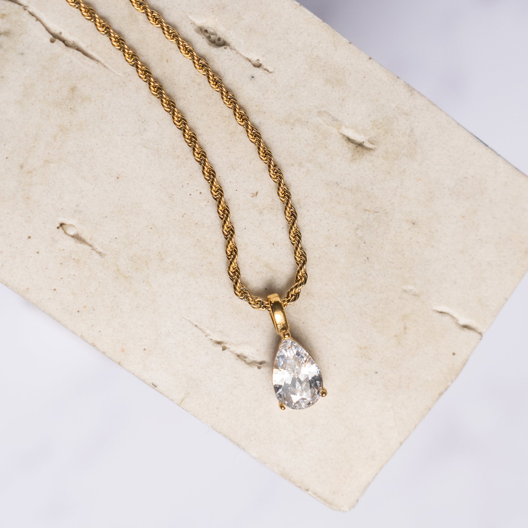 A gold-colored necklace with a clear teard-shaped zirconia pendant displayed on a beige background.