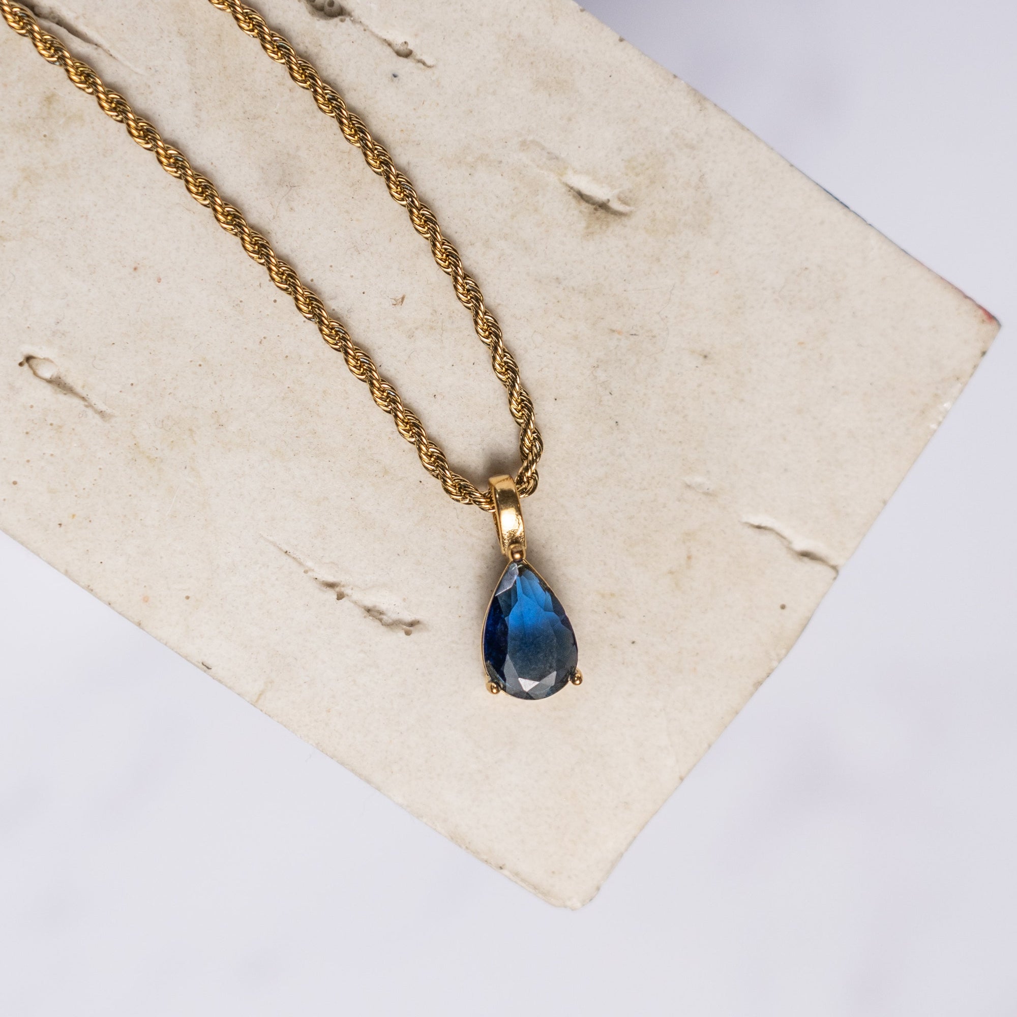 A blue sapphire necklace with a gold chain, displayed on a beige background.