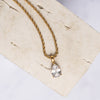 A gold-colored necklace with a clear teard-shaped zirconia pendant displayed on a beige background.