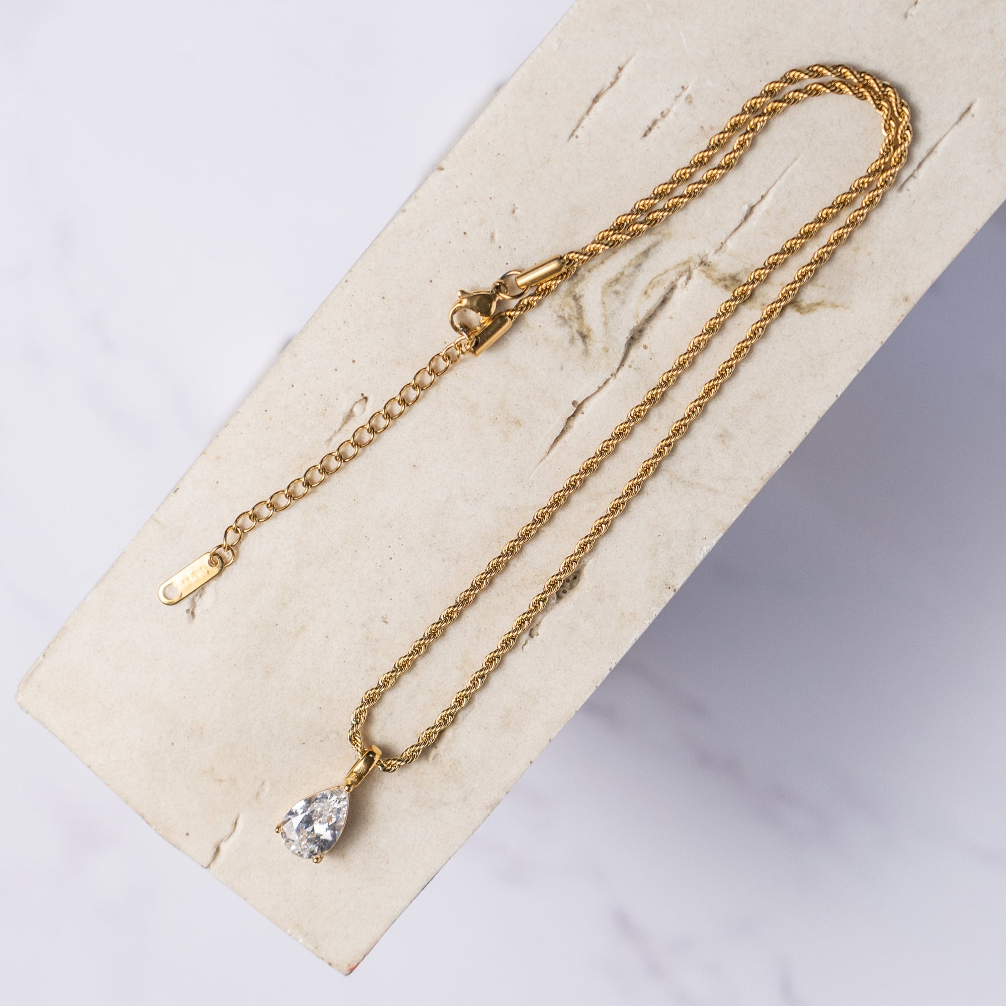 A gold-colored necklace with a clear teard-shaped zirconia pendant displayed on a beige background.