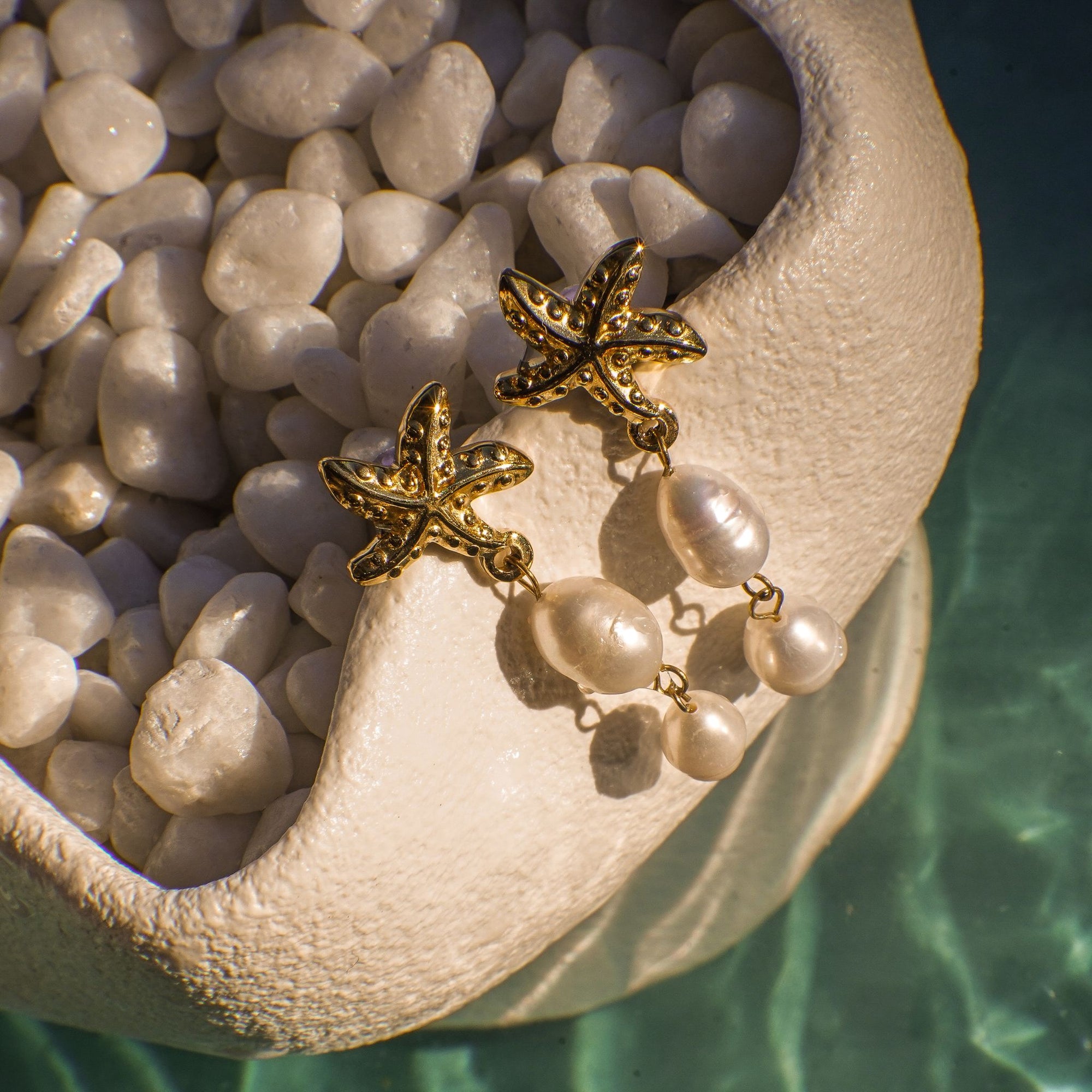 Gold starfish earrings with pearl drops on a shell against a pebble background