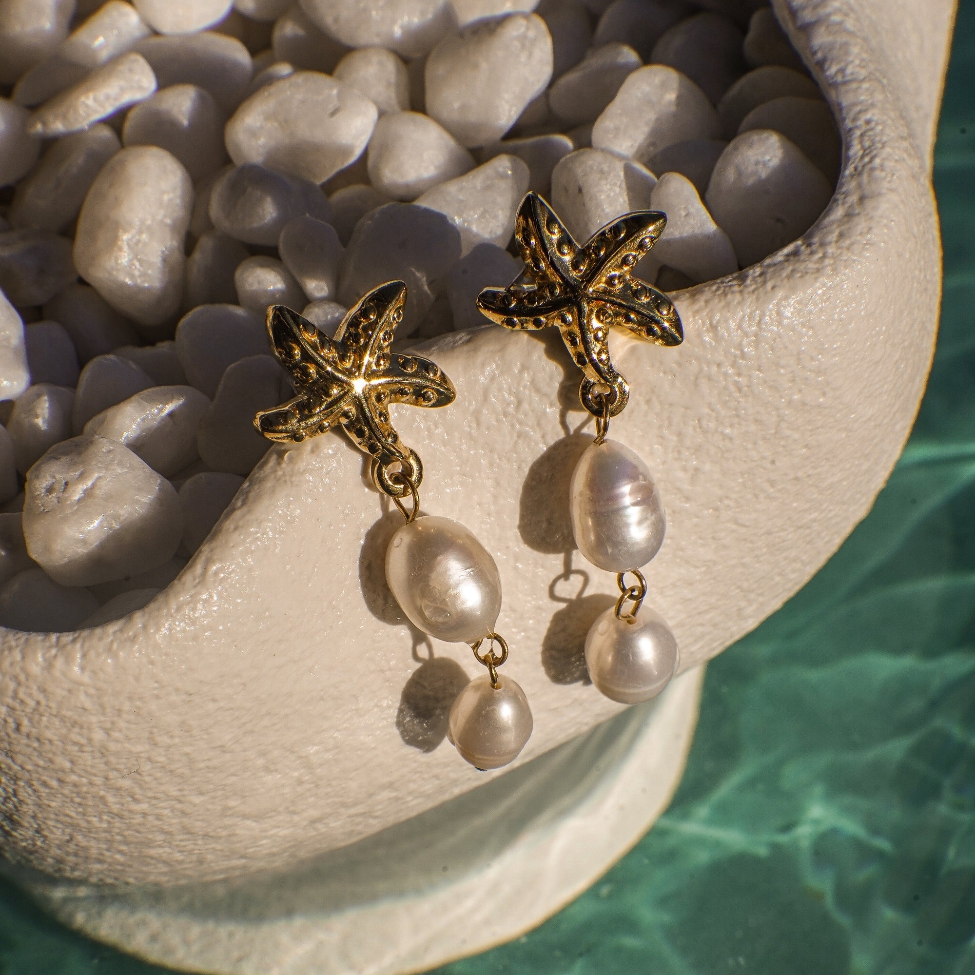 Gold starfish earrings with pearl drops on a stone surface near water.