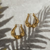 A pair of gold drop hoop earrings with a textured design, displayed on a grey surface with a shadow of a plant leaf overhead.