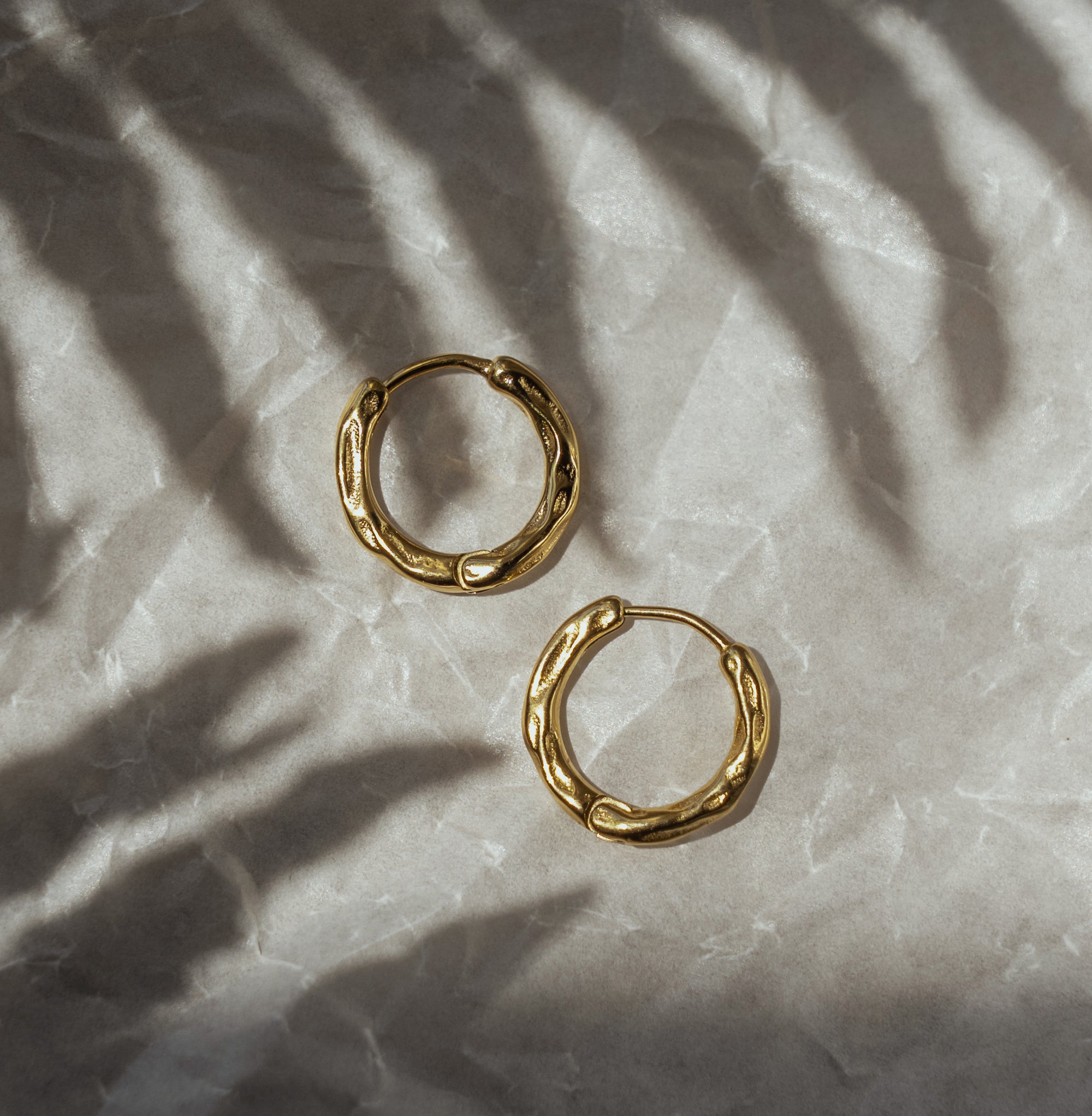 A pair of gold-colored hoop earrings on a textured background with a shadow cast over them.