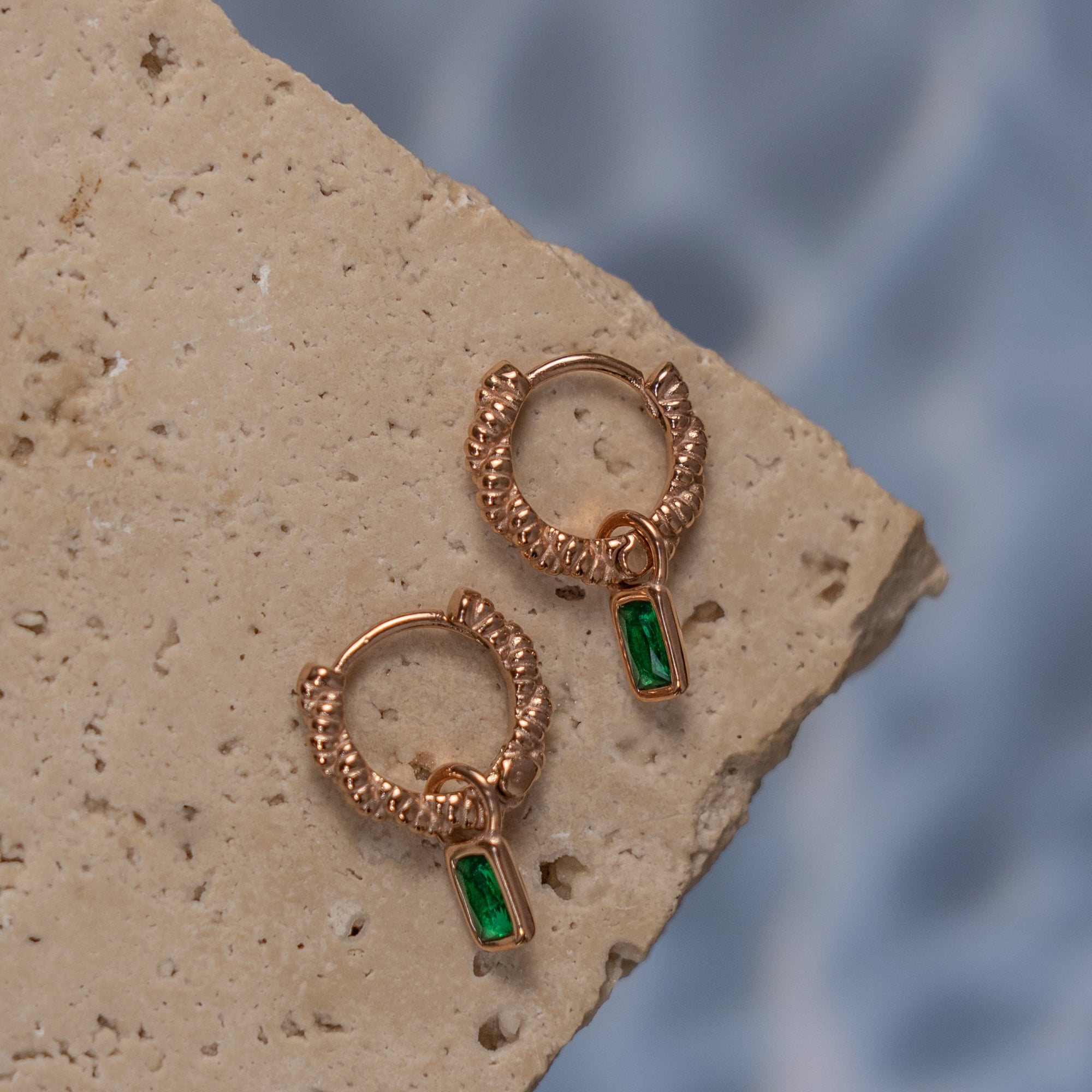 A pair of rose gold earrings with Emerald drop pendants on a textured surface.