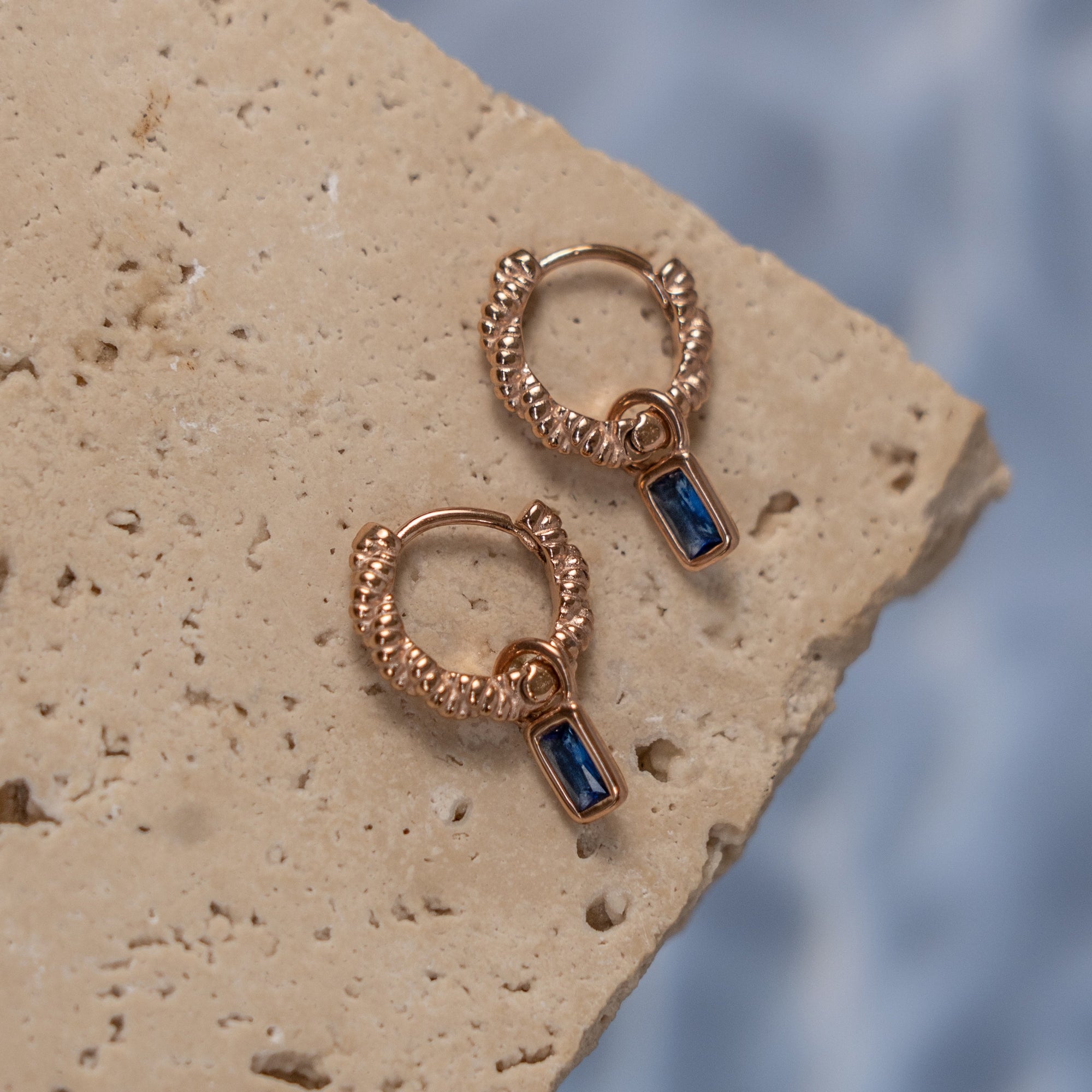 A pair of rose gold earrings with sapphire drops on a textured surface