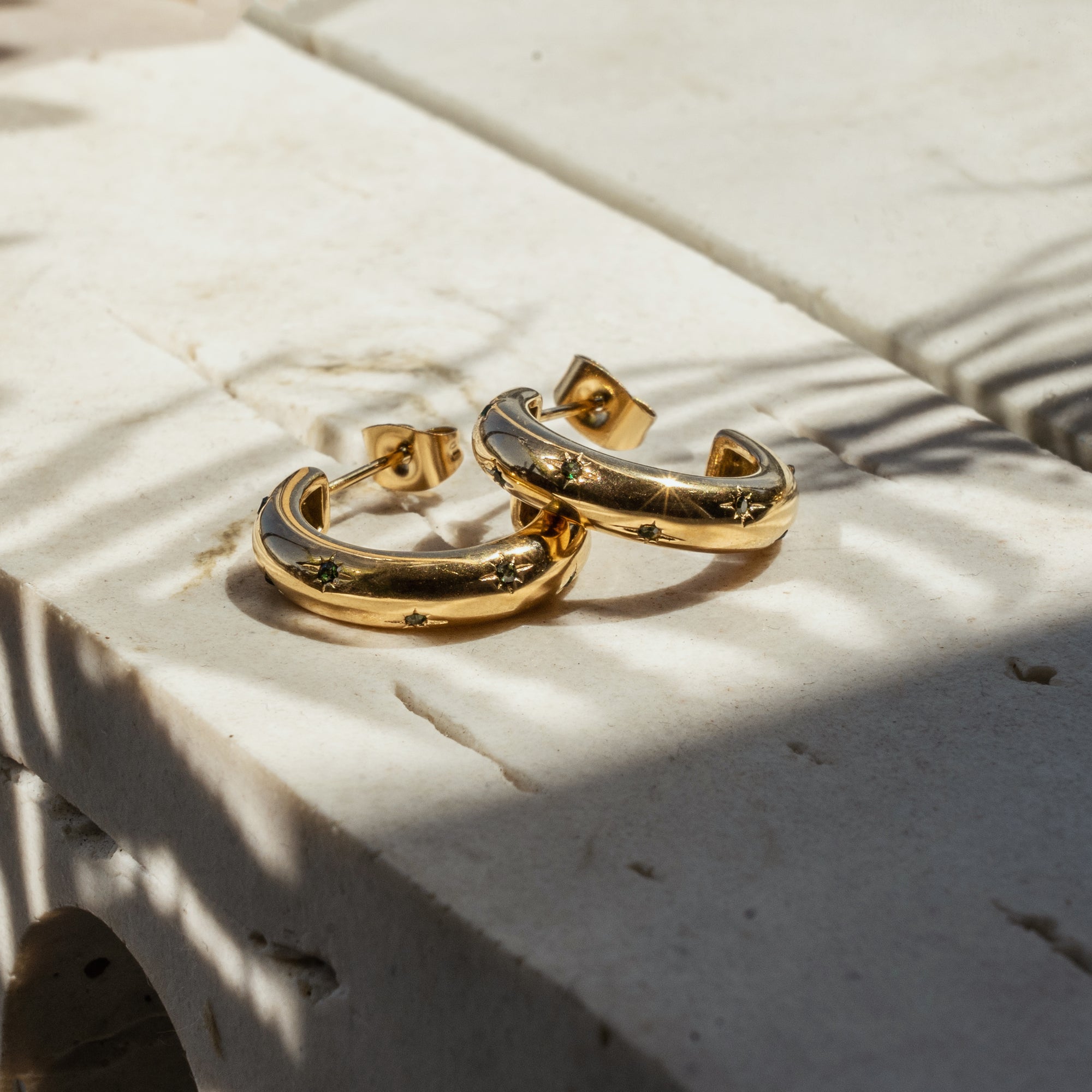 A pair of gold-colored hoop earrings with emerald star zirconia embedded on a stone surface.