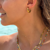 A model wearing a gold hoop earring with emerald accents made out of zirconia paired with other gold, colorful necklaces